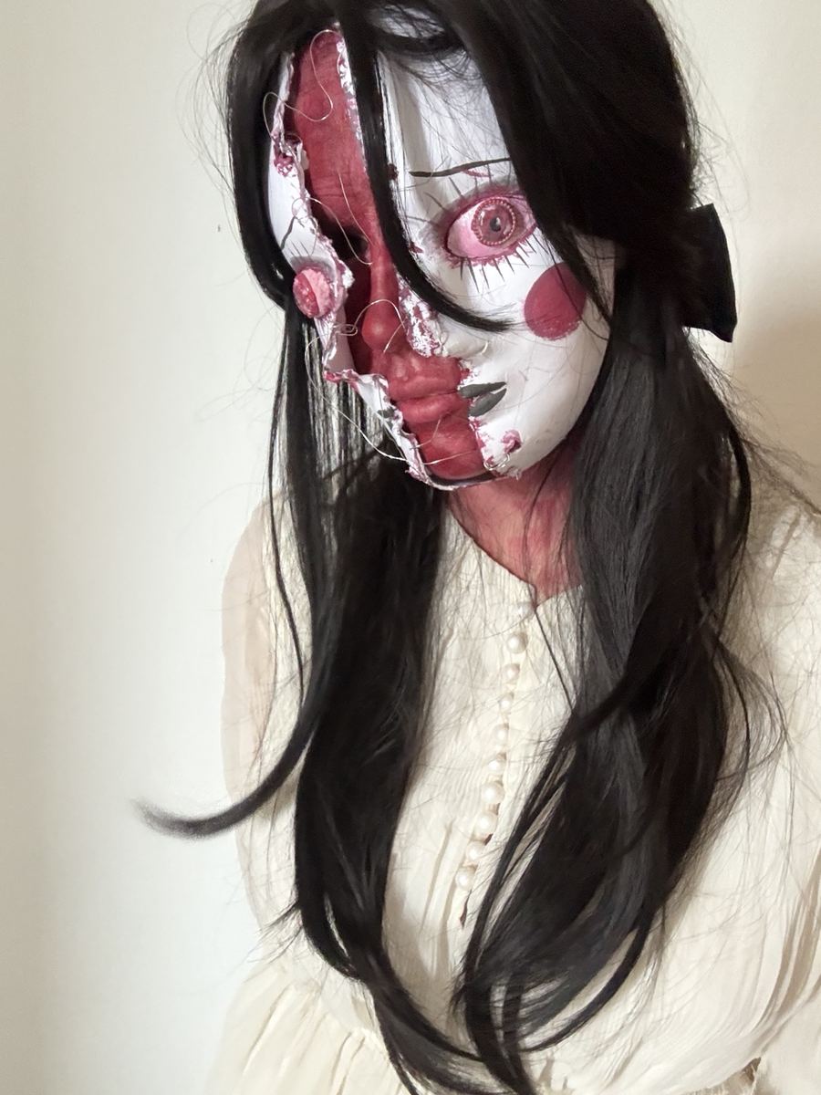 Porcelain doll character: painted face splitting open over raw tissue, stitched with thread