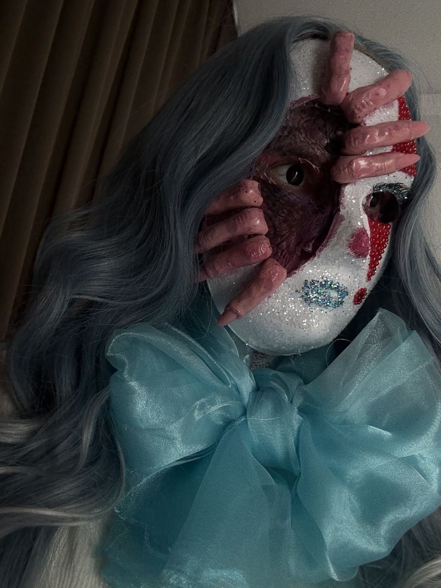 Character with a glittered half mask lifted away from raw tissue beneath, pink sculpted fingers