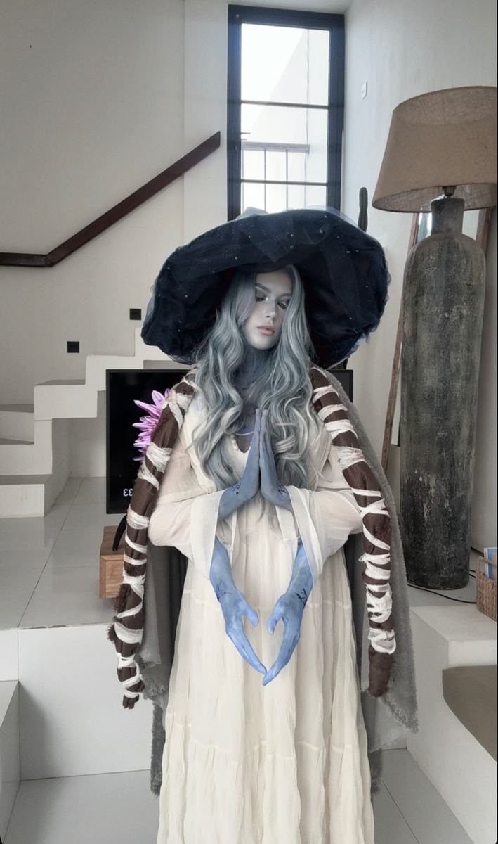 Sorceress cosplay in a pale gown with blue sculpted hands and a wide veiled hat